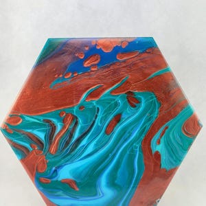 Può includere: Piastrella decorativa esagonale con un design astratto. La piastrella presenta una miscela di colori, tra cui verde acqua, rame e blu, creando un effetto fluido e marmorizzato. La piastrella è esposta su un supporto trasparente.