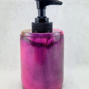May include: A cylindrical soap dispenser with a black pump top. The body of the dispenser is a gradient of pink and purple with a glittery finish. The dispenser is designed to hold liquid soap.