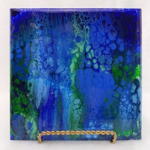 May include: Square ceramic tile with an abstract design in shades of blue and green. The artwork features a fluid, cell-like pattern, with a glossy finish. The tile is displayed on a gold-colored metal stand.