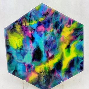 May include: A hexagonal art piece with a vibrant, abstract design. The artwork features a blend of colors, including blue, yellow, pink, and purple, creating a dynamic and eye-catching visual effect. The piece is displayed on a clear stand.