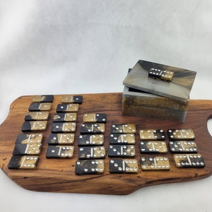 May include: A set of black and gold dominoes with white dots on a wooden board. The dominoes are in a box with a lid.