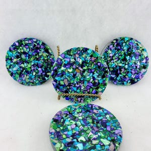 May include: Four round coasters with a mosaic design in shades of blue, green, and purple. One coaster is displayed on a small gold-colored metal stand. The coasters are made of a shiny material.