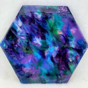 May include: Hexagonal coaster with an abstract design. The coaster displays a swirling mix of colours, including deep purples, blues, greens, and pink, creating a galaxy-like effect. The edges are smooth and the surface is glossy.
