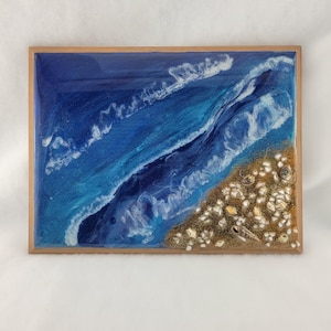 May include: Resin artwork of a beach scene in a wooden frame. The image shows a blue ocean with white waves, a sandy shore with seashells, and a natural wood frame. The artwork evokes a coastal theme.