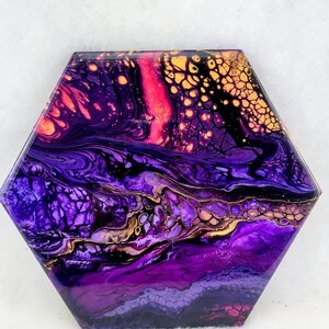 May include: Hexagonal abstract artwork with a fluid design. The piece features a blend of purple, gold, and pink hues, creating a vibrant visual effect. Likely a decorative item or a coaster, with dimensions in centimetres.