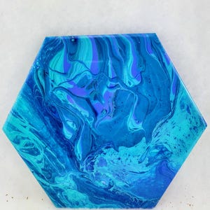 Puede incluir: Pieza de arte hexagonal con un diseño abstracto y fluido. La obra presenta una mezcla de azules y turquesas, creando un efecto marmolado. Los colores se arremolinan juntos, dando la impresión de agua u olas. Probablemente sea un posavasos o una baldosa decorativa.