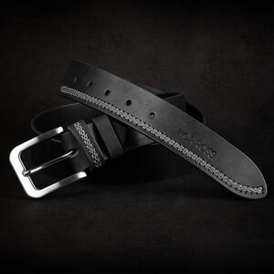 May include: A black leather belt with a silver buckle. The belt has white stitching along the edges and the brand name "TORROSS" is embossed on the belt. The belt is rolled up on a dark background.