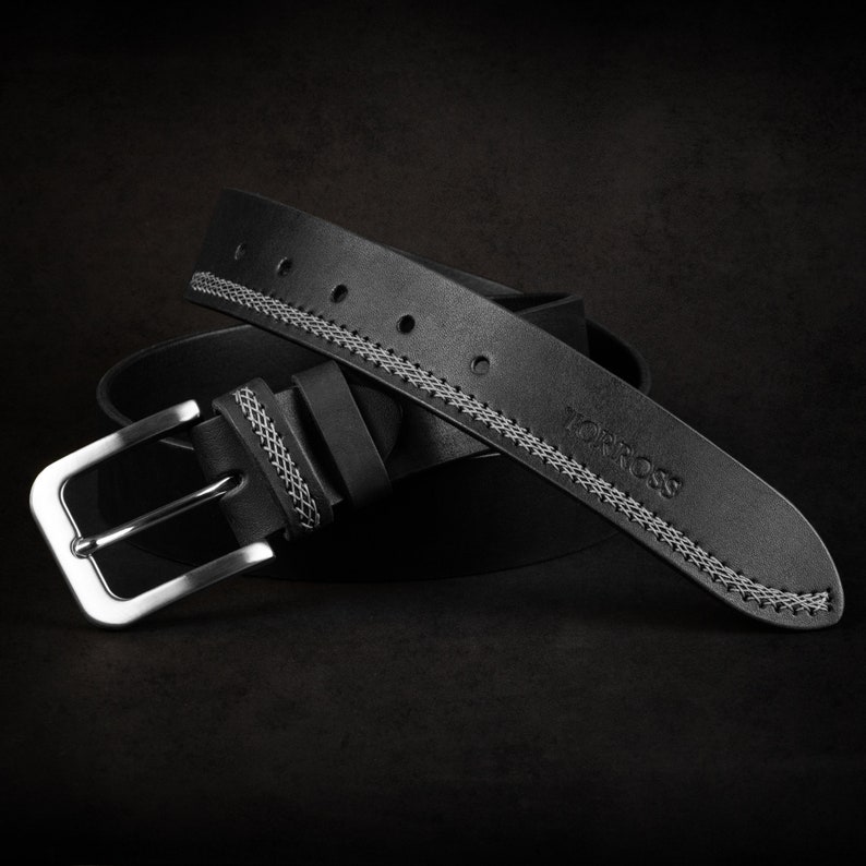 May include: A black leather belt with a silver buckle. The belt has white stitching along the edges and the brand name "TORROSS" is embossed on the belt. The belt is rolled up on a dark background.