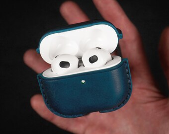 Puede incluir: Una funda de cuero azul oscuro para auriculares inalámbricos. La funda está abierta, revelando dos auriculares blancos con detalles negros.