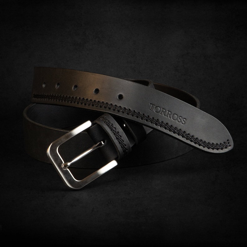 May include: A black leather belt with a silver buckle. The belt has decorative stitching along the edges and the word "TORROSS" is embossed on the belt. The belt is shown against a dark background.