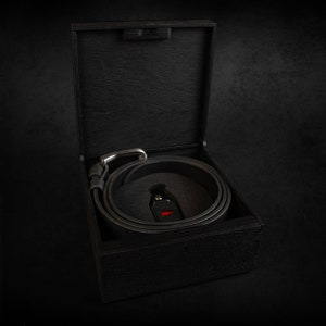 May include: A black leather belt with a silver buckle is coiled inside a dark wooden box. The box is open, revealing the belt nestled in a black foam insert. A small, red-accented tag is visible.