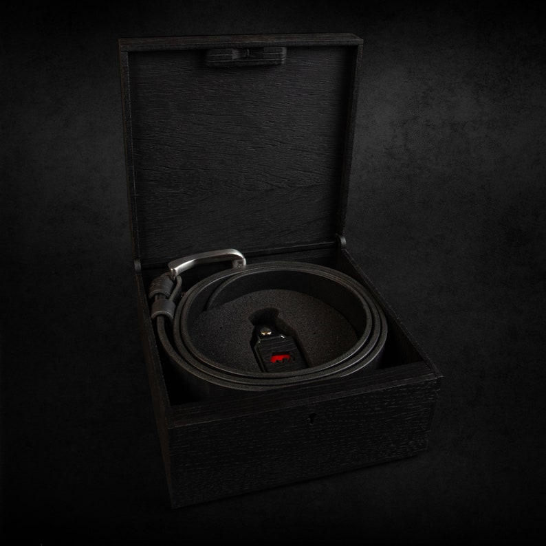 May include: A black leather belt with a silver buckle is coiled inside a dark wooden box. The box is open, revealing the belt nestled in a black foam insert. A small, red-accented tag is visible.