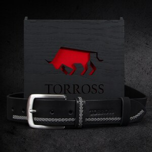 May include: Black leather belt with a silver buckle and white stitching detail. The belt is displayed in front of a black wooden box with a red bull logo and the word "TORROSS".