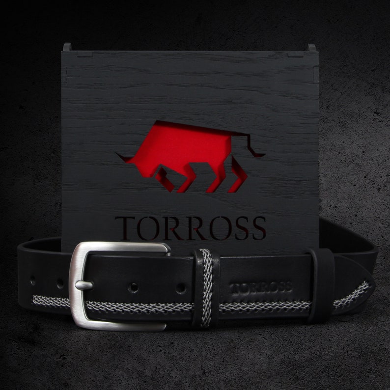 May include: Black leather belt with a silver buckle and white stitching detail. The belt is displayed in front of a black wooden box with a red bull logo and the word "TORROSS".