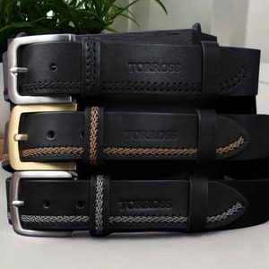 May include: Three black leather belts with silver, gold, and silver buckles. Each belt has decorative stitching and the brand name "TORROSS" embossed on the leather. The belts are stacked, showcasing their different buckle styles and stitching details.