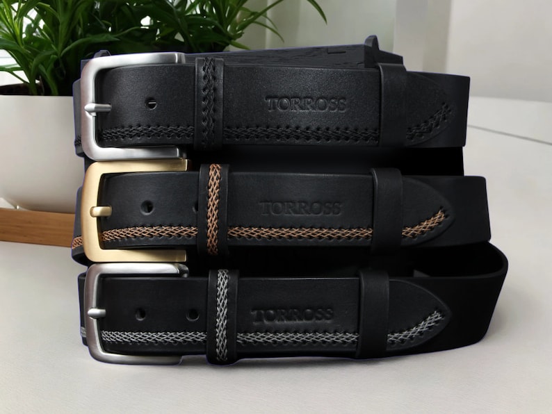 May include: Three black leather belts with silver, gold, and silver buckles. Each belt has decorative stitching and the brand name "TORROSS" embossed on the leather. The belts are stacked, showcasing their different buckle styles and stitching details.