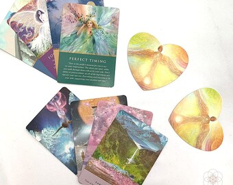 May include: A set of angel oracle cards with colorful illustrations and text. The cards are arranged in a fan shape on a white surface. Two heart-shaped cards with angel imagery are also visible. The text on one card reads "Perfect Timing" and another card reads "Earth Pulsing".