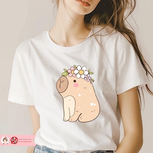 May include: White t-shirt featuring a cartoon illustration of a capybara wearing a flower crown. The capybara is light brown with a light pink nose and small details. The shirt has the text "Hand Drawn 100% No AI Generated."