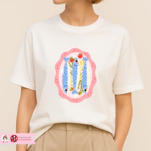 Puede incluir: Camiseta blanca con una colorida ilustraci&oacute;n de tres peces azules sobre un plato rosa, adornada con hierbas, lim&oacute;n y un tomate. El dise&ntilde;o tiene una est&eacute;tica dibujada a mano. La camiseta es de material suave.