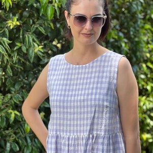 May include: A woman wearing a sleeveless, light purple and white gingham dress. She is standing in front of a green leafy background and wearing sunglasses.
