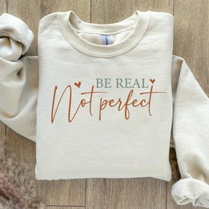 Op de afbeelding: Cr&egrave;mekleurige sweatshirt met de woorden "BE REAL, Not perfect" in roest- en saliegroen. De sweatshirt heeft een ronde hals en lange mouwen. De tekst is handgeschreven.