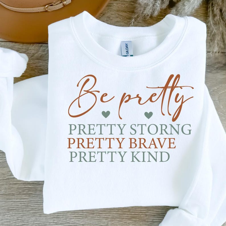 Op de afbeelding: Witte sweatshirt met de tekst "Be pretty" in bruin script. Daaronder staan de woorden "PRETTY STRONG, PRETTY BRAVE, PRETTY KIND" in bruin en groen. De sweatshirt is opgevouwen.