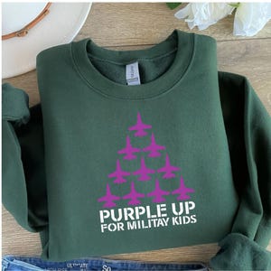 May include: A dark green sweatshirt with a white graphic of a purple airplane formation and the text "Purple Up for Military Kids".