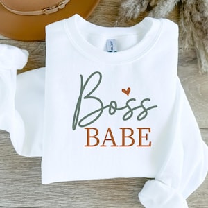 Op de afbeelding: Witte sweatshirt met de woorden "Boss Babe" in groene en bruine letters. Een klein rood hart staat boven het woord "Boss". De sweatshirt is opgevouwen, met de mouwen erin.