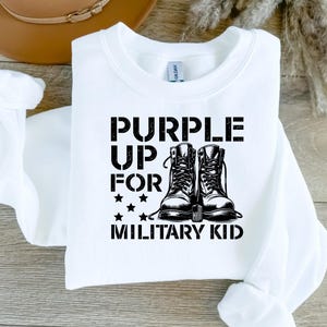 Pu&ograve; includere: Felpa bianca con testo nero che recita "Purple Up For Military Kid" e una grafica di due stivali militari neri con stelle.