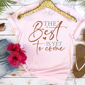 Op de afbeelding: Lichtroze t-shirt met de tekst "The Best is Yet to Come" in bruine en groene letters. Het shirt wordt gepresenteerd op een gouden hanger, met een zonnebril en bloemen op de achtergrond.