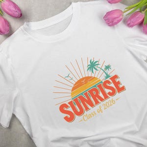 以下が含まれることがあります： レトロな日の出のデザインが施された白いTシャツ。ヤシの木、鳥、オレンジ色の「SUNRISE」の文字が描かれています。「Class of 2026」の文字が下にあります。