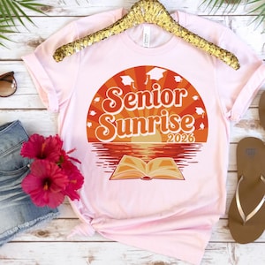 以下が含まれることがあります： オレンジと白で「Senior Sunrise 2026」の文字が描かれたグラフィックデザインのライトピンクのTシャツ。 デザインには、開いた本と卒業キャップのある夕日のシーンが含まれています。