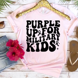 Pu&ograve; includere: Maglietta rosa con testo nero che recita "Purple Up For Military Kids" con un simbolo di nastro viola.