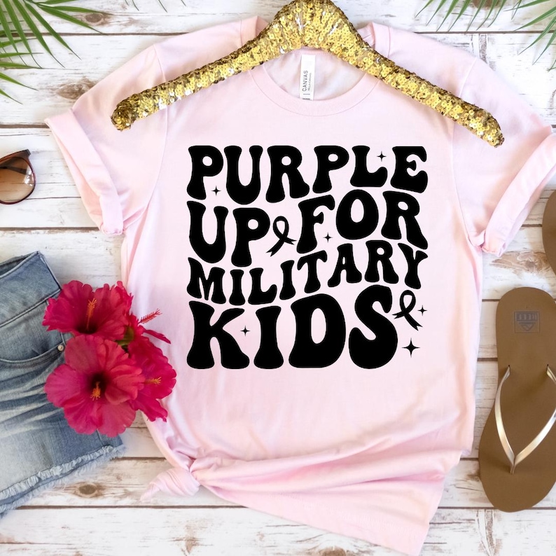 Pu&ograve; includere: Maglietta rosa con testo nero che recita "Purple Up For Military Kids" con un simbolo di nastro viola.