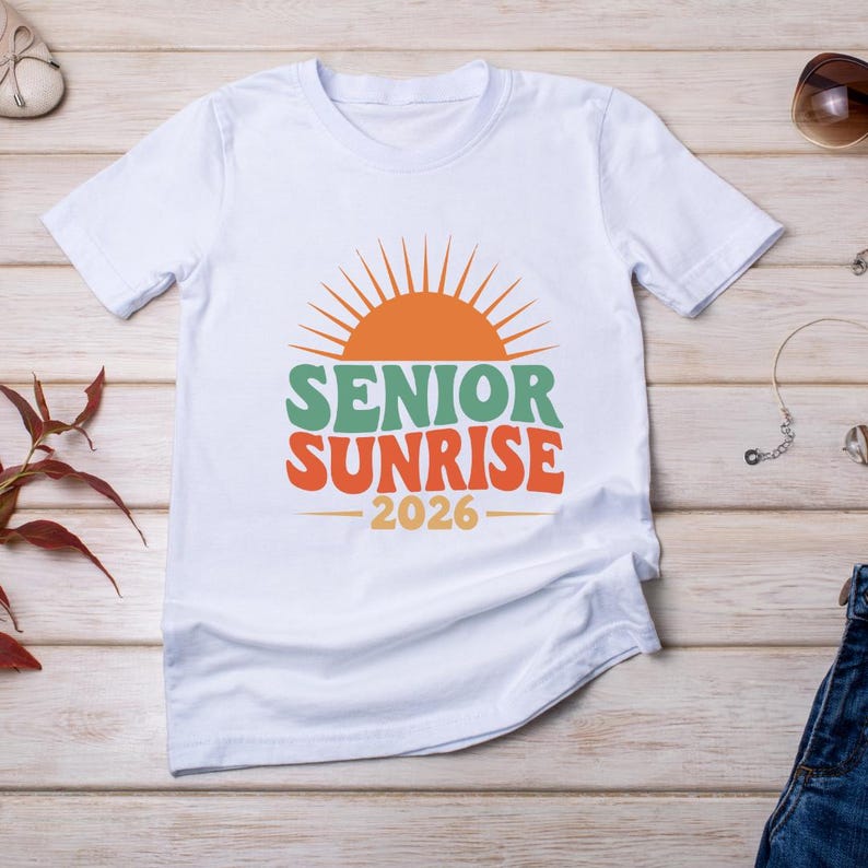 以下が含まれることがあります： カラフルなグラフィックが施された白いTシャツ。デザインは、緑とオレンジ色の「SENIOR SUNRISE」という言葉の上にオレンジ色のサンバースト、その下に「2026」が描かれています。柔らかい素材で作られたシャツです。