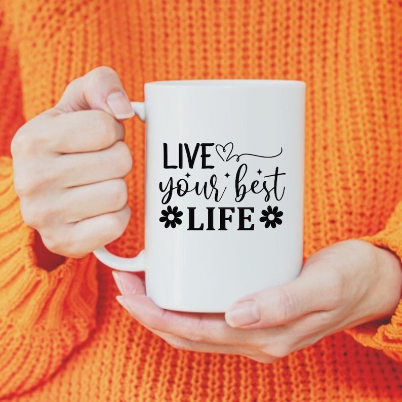 K&ouml;nnte beinhalten: Eine wei&szlig;e Keramiktasse mit dem Schriftzug "LIVE your best LIFE" in schwarzer Schrift und floralen Akzenten. Die Tasse wird von einer Person gehalten, die einen orangefarbenen Pullover tr&auml;gt. Die Tasse hat eine Standardgr&ouml;&szlig;e f&uuml;r Kaffee oder Tee.