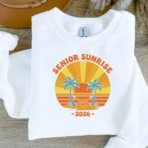 以下が含まれることがあります： オレンジと黄色で「Senior Sunrise」と書かれたカラフルなグラフィックが特徴の白いスウェットシャツ。デザインには太陽、ヤシの木、ハンモックが含まれており、下に「2026」の年号があります。スウェットシャツは柔らかい素材で作られています。