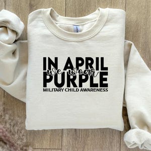 Pu&ograve; includere: Una felpa color crema con il testo "In April we wear purple" e "Military Child Awareness" stampato in nero.