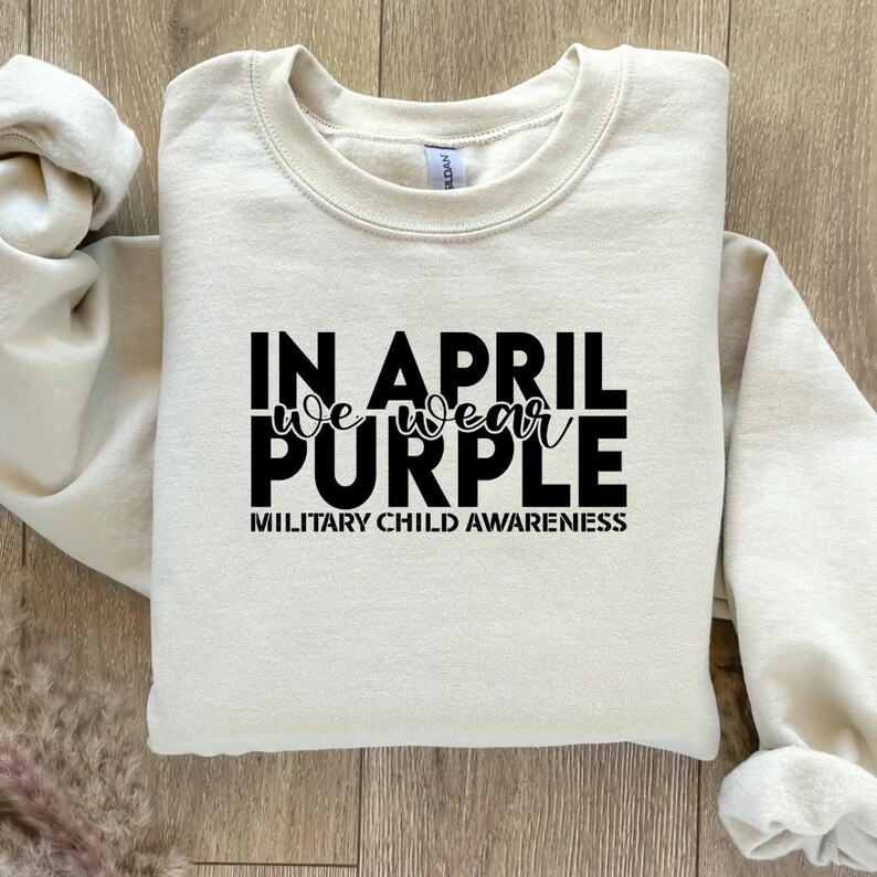 Pu&ograve; includere: Una felpa color crema con il testo "In April we wear purple" e "Military Child Awareness" stampato in nero.