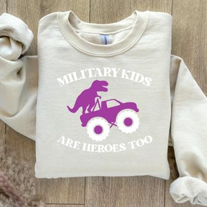 May include: A beige sweatshirt with the text "Military Kids Are Heroes Too" in white. A purple dinosaur is riding on top of a purple monster truck.