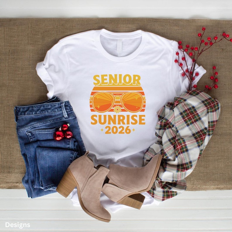 以下が含まれることがあります： オレンジと黄色の「SENIOR SUNRISE +2026+」グラフィックがプリントされた白いTシャツ。青いジーンズ、タン色のアンクルブーツ、チェック柄のシャツと組み合わせています。赤い装飾用のベリーも含まれています。
