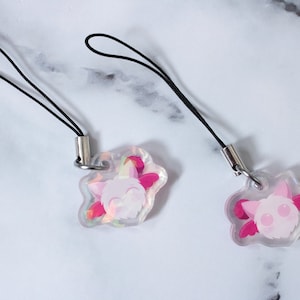 May include: Two clear acrylic charms with black cords. Each charm features a pink cartoon character with pink wings and a smiling face. The charms are attached to silver-colored metal loops.