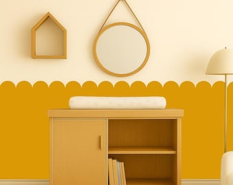 Op de afbeelding: Een kinderkamer met een houten commode, een ronde spiegel en een huisvormig rek. De muur is geschilderd met een mosterdgeel geschulpd ontwerp. Een witte aankleedkussen ligt op de tafel. Een vloerlamp staat op de achtergrond.