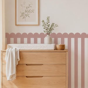 以下が含まれることがあります： 3段の引き出しがある明るい木製のチェスト。上には白いおむつ替えマットと白いタオルが置かれています。背面の壁はくすんだピンクと白の縦縞模様で、上部はスカラップ状の縁取りになっており、その上には植物の絵が額装されて飾られています。