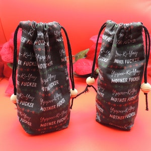 Puede incluir: Dos bolsas negras con cordón con un patrón de texto blanco repetido que dice "Yippee-Ki-Yay Mother Fucker". Las bolsas tienen cordones negros y detalles de cuentas de madera. El fondo es rojo brillante.