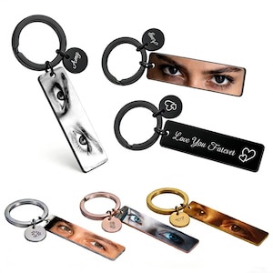 May include: A collection of rectangular metal keychains in black, silver, rose gold, and gold finishes. Each keychain features a split ring, a small circular charm with engraved icons or names, and a rectangular tag displaying eye portraits.