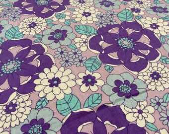 Op de afbeelding: Paarse, witte en blauwe bloemenstof met een retro-ontwerp. De stof heeft grote paarse bloemen met witte centra en kleinere blauwe bloemen met witte centra. De achtergrond is lichtpaars.