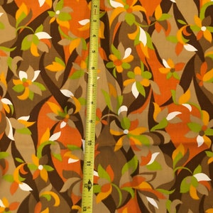 Op de afbeelding: Een bruine stof met een herhalend patroon van oranje, groene en witte gestileerde bloemen. De stof wordt gemeten met een gele meetlint.