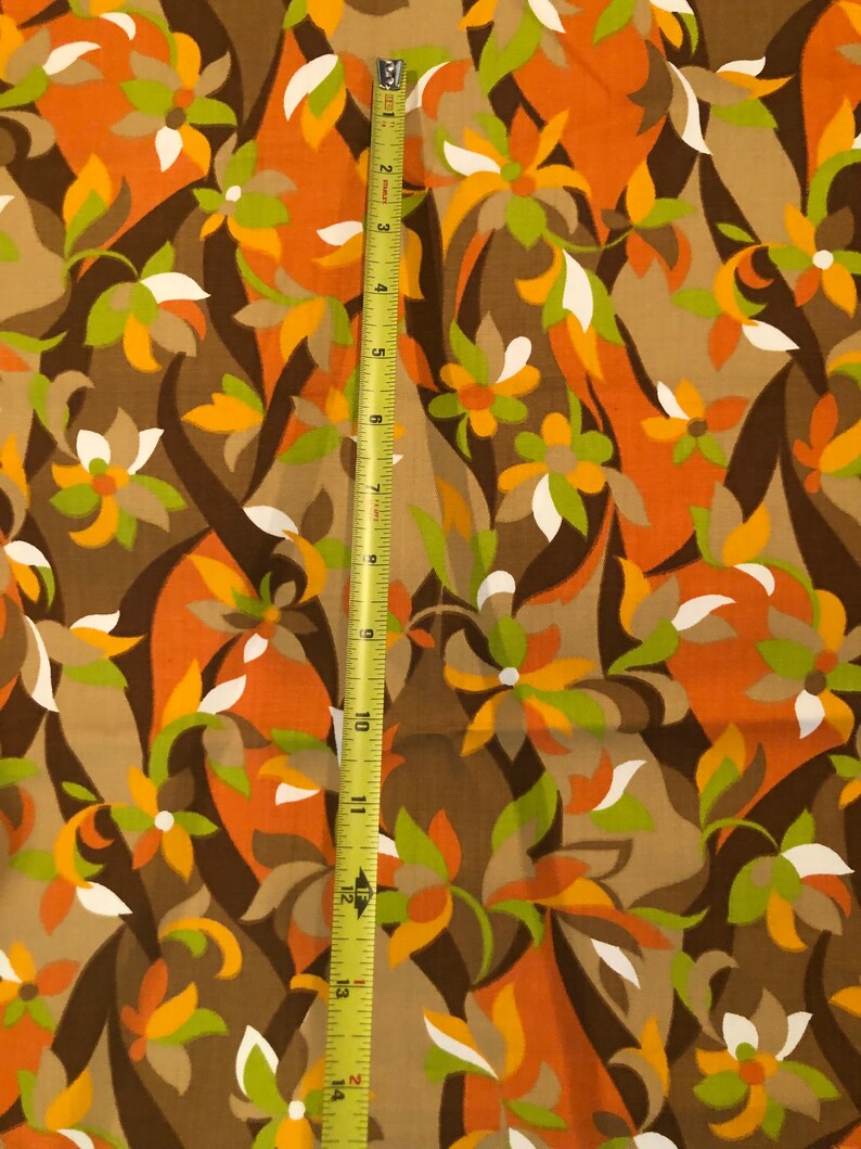 Op de afbeelding: Een bruine stof met een herhalend patroon van oranje, groene en witte gestileerde bloemen. De stof wordt gemeten met een gele meetlint.