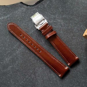 Peut inclure: Un bracelet de montre en cuir marron lisse doté d'une boucle déployante en métal argenté brillant. Le bracelet comprend deux passants en cuir assortis et une série de trous perforés pour le réglage, avec des coutures visibles sur les bords.